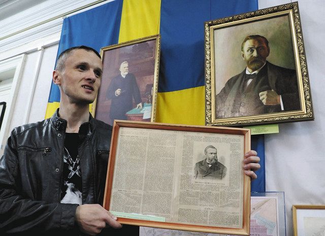 アルフレド・ノーベルの肖像画（右）の前で、ロシア紙に載った兄リュドビクの死亡記事を掲げるリャボイ館長＝ロシア西部ヤロスラブリ州ルイビンスクで