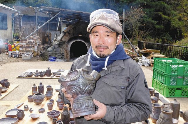 穴窯（後方）で焼いた自身の作品を持つ薬王寺さん＝いずれも横須賀市で 